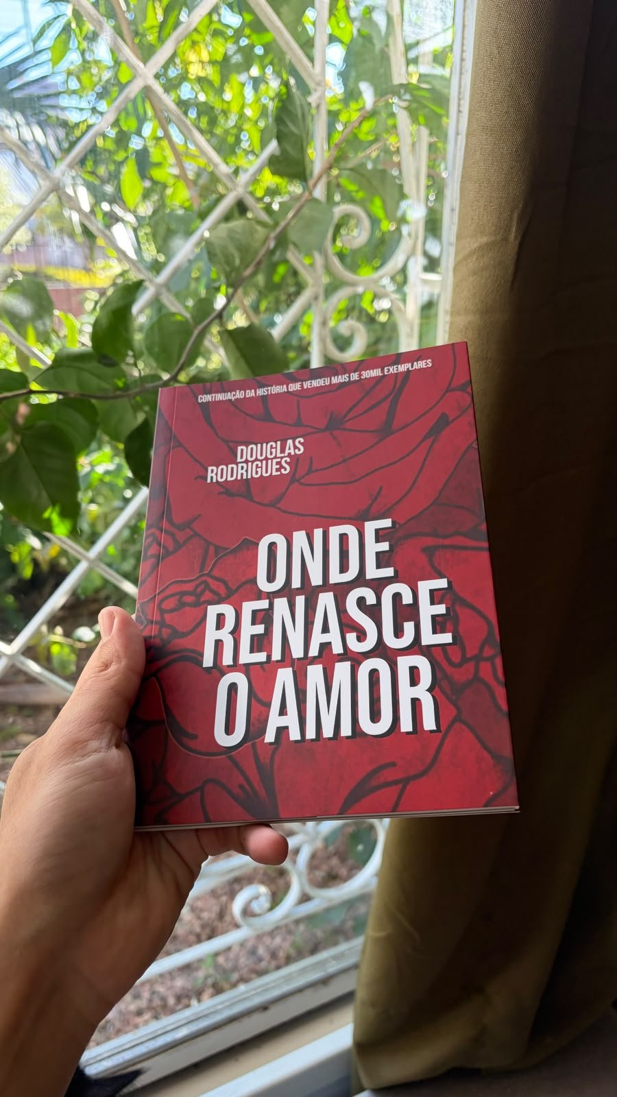 Onde Renasce o Amor sendo segurado diante de uma janela com folhagem ao fundo
